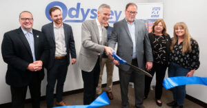 Odyssey employees standing next to each other as Hans cuts a blue ribbon in half with giant scissors